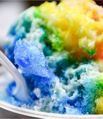 A close up of a spoon dipping into a rainbow colored serving of Hawaiian shave ice.