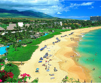 An overhead view of a populated beach in Hawaii. The composition shows the beach is backed by a resort and green mountains.