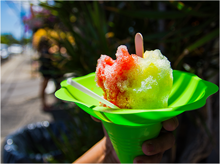 A hand holds a serving of shaved ice in the shade. Two flavors have been combined and sit in a plastic cone.