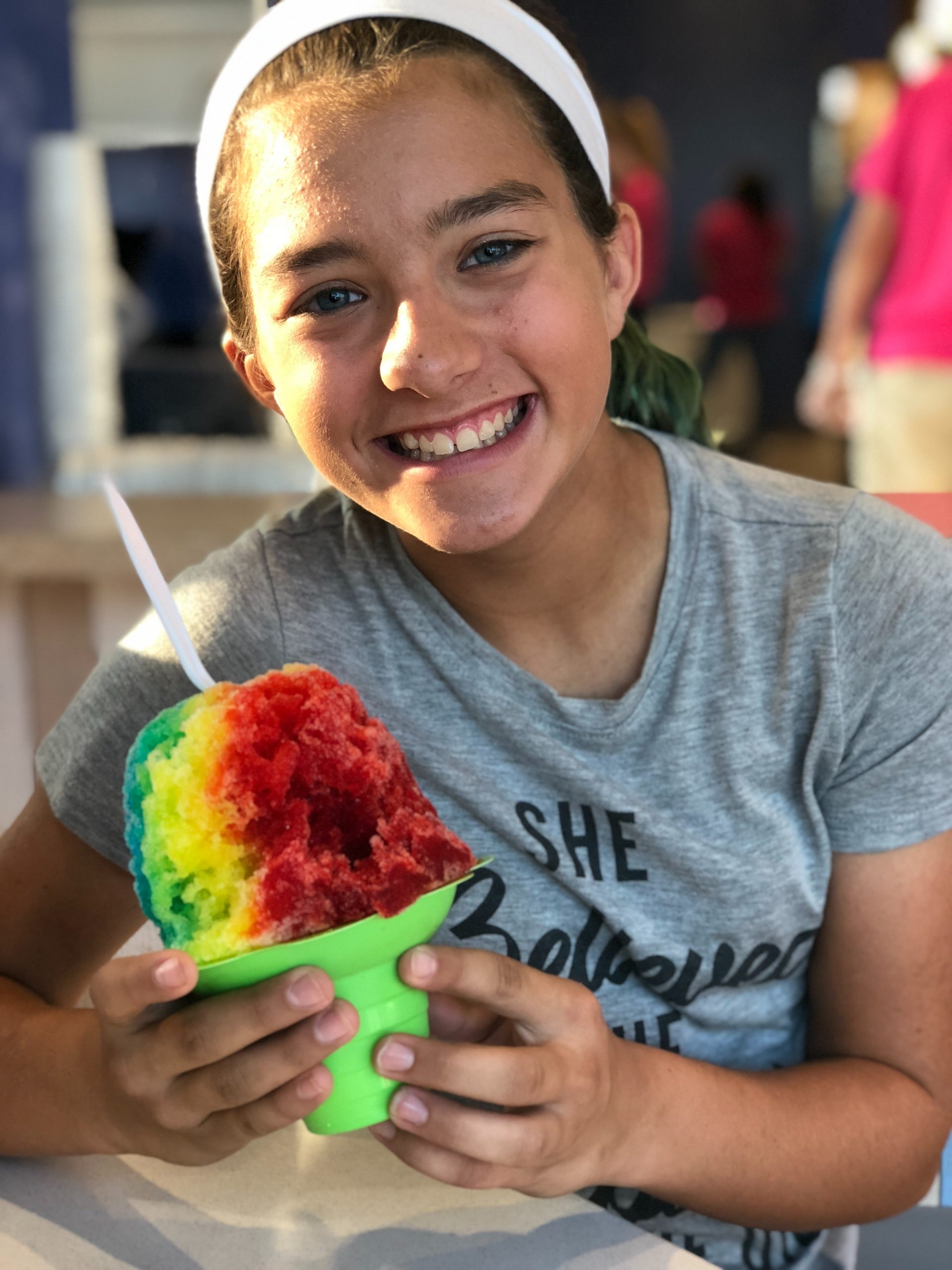 A young girl poses for a photo while holding a large serving of shaved ice. The flavors are in a rainbow pattern.