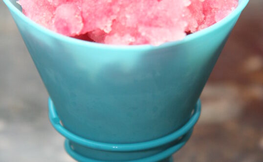 Reddish pink Hawaiian shave ice is served in a plastic cone.